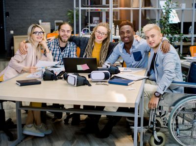 Inclusion In Work, Business Team Concept. Handsome Male Coworker On Wheelchair With His Creative Business Colleagues In Meeting Room At Creative Office, Smiling, Looking At Camera, Hugging Each Other