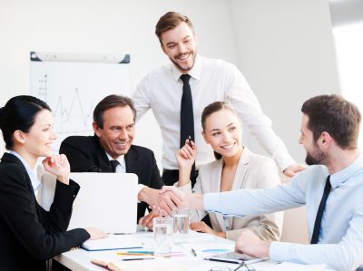 Welcome on board! Group of confident business people in formalwear sitting at the table together and smiling while two men handshaking